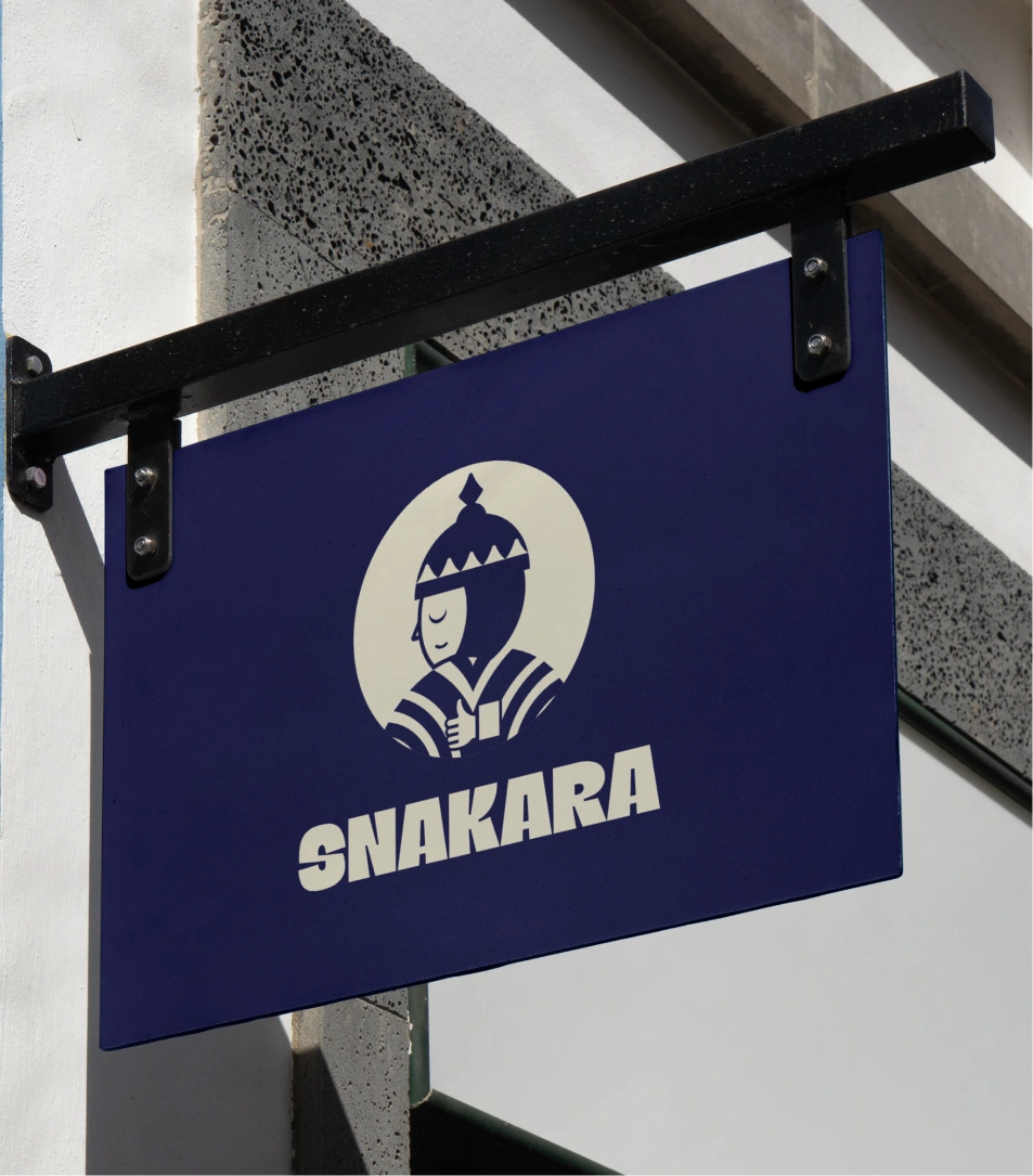 This image displays **Snakara’s outdoor brand signage**, featuring its signature mascot and bold logotype. The signage is mounted on a black metal bracket against a modern white and grey building facade, exuding a crisp and urban aesthetic. The deep navy-blue background contrasts elegantly with the ivory illustration of the *Snakara Samurai* — a stylized guardian figure that embodies the brand’s spirit of mindful strength and everyday balance. The thick, confident typography below enhances visibility and impact, making the logo instantly recognizable. This visual captures Snakara’s essence as a contemporary Indian snacking brand that blends tradition, character, and modern design sensibility in every detail.
