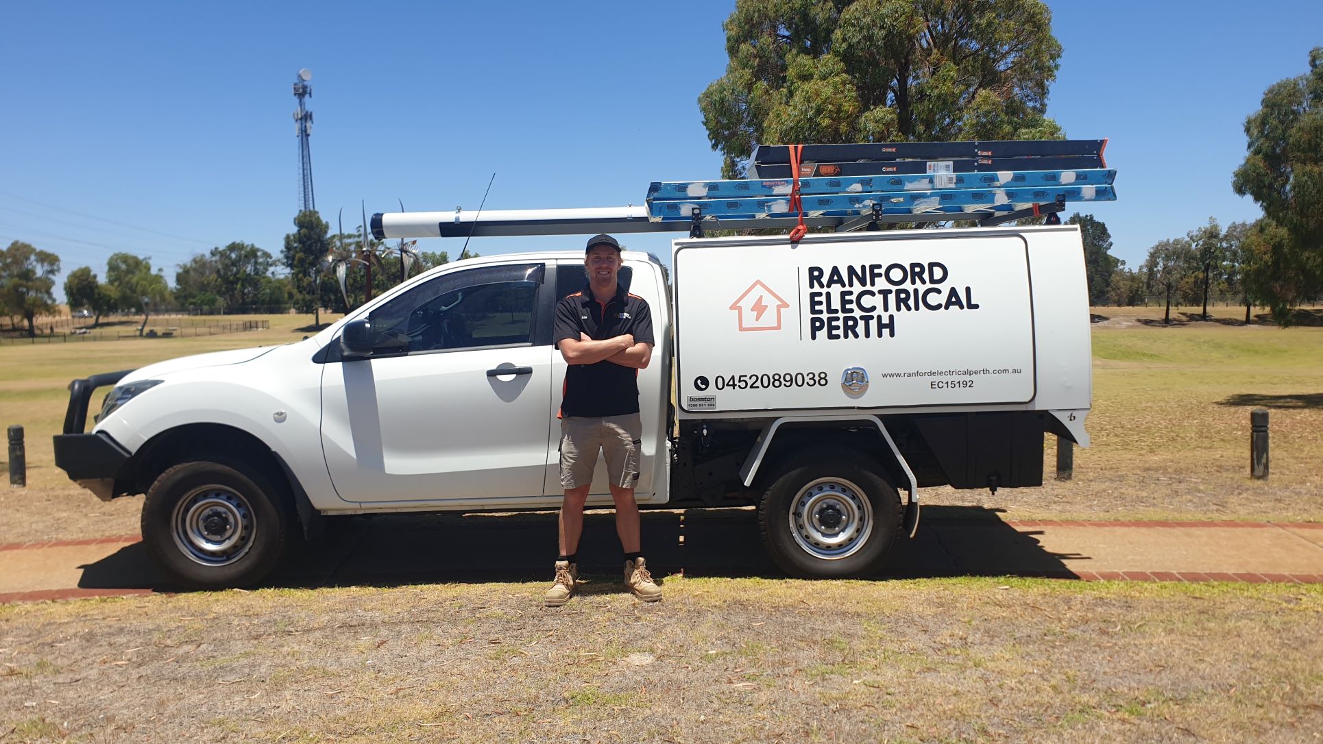 Ash Ranford in black shirt and shorts standing with arms crossed beside a white electrical service truck with ladders on top.