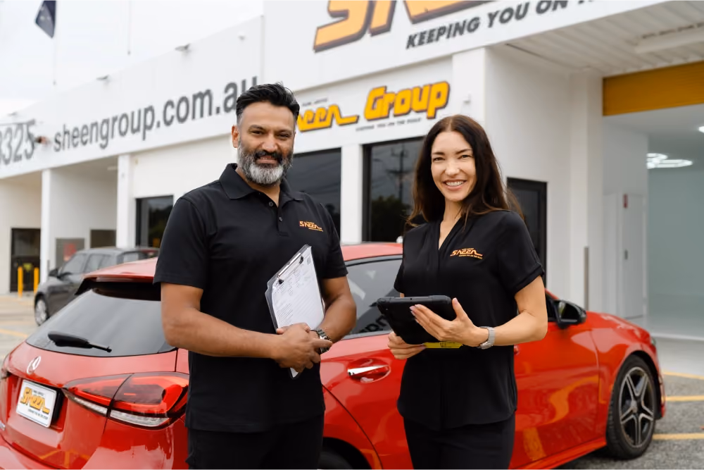 Two Sheen Group staff members smiling outside a Sheen Group service centre, standing in front of a red car while holding a clipboard and tablet.