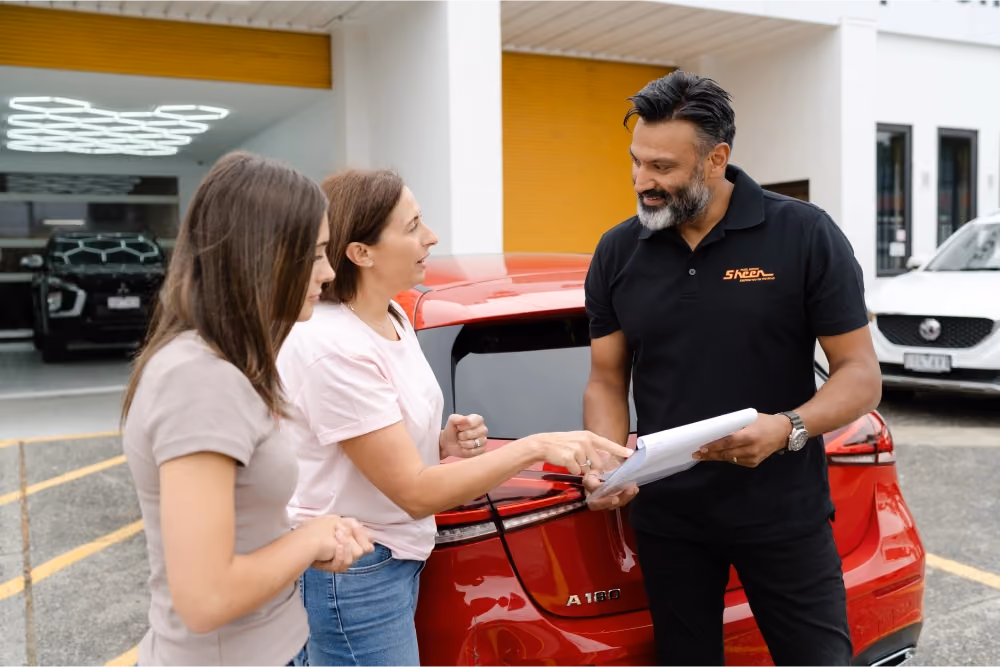 Two women discussing paperwork with a Sheen Group staff member next to a red car outside the repair centre.