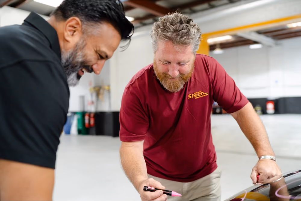 Two Sheen Group team members inside a workshop, smiling and discussing a vehicle repair while one holds a pink marker.