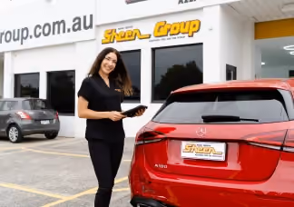 Sheen Group staff member smiling while holding a tablet, standing beside a red car outside a Sheen service centre.