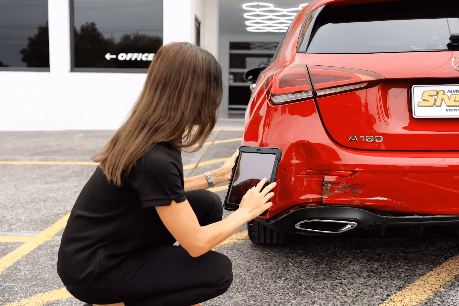 Sheen staff member kneeling beside a red Mercedes A180, using a tablet to photograph rear bumper damage in a repair centre car park.