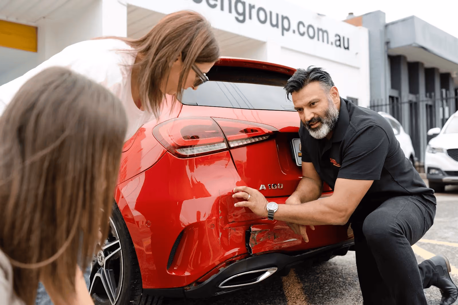 Panel technician inspecting rear bumper damage on a red car while speaking with two customers outside a Sheen repair centre.