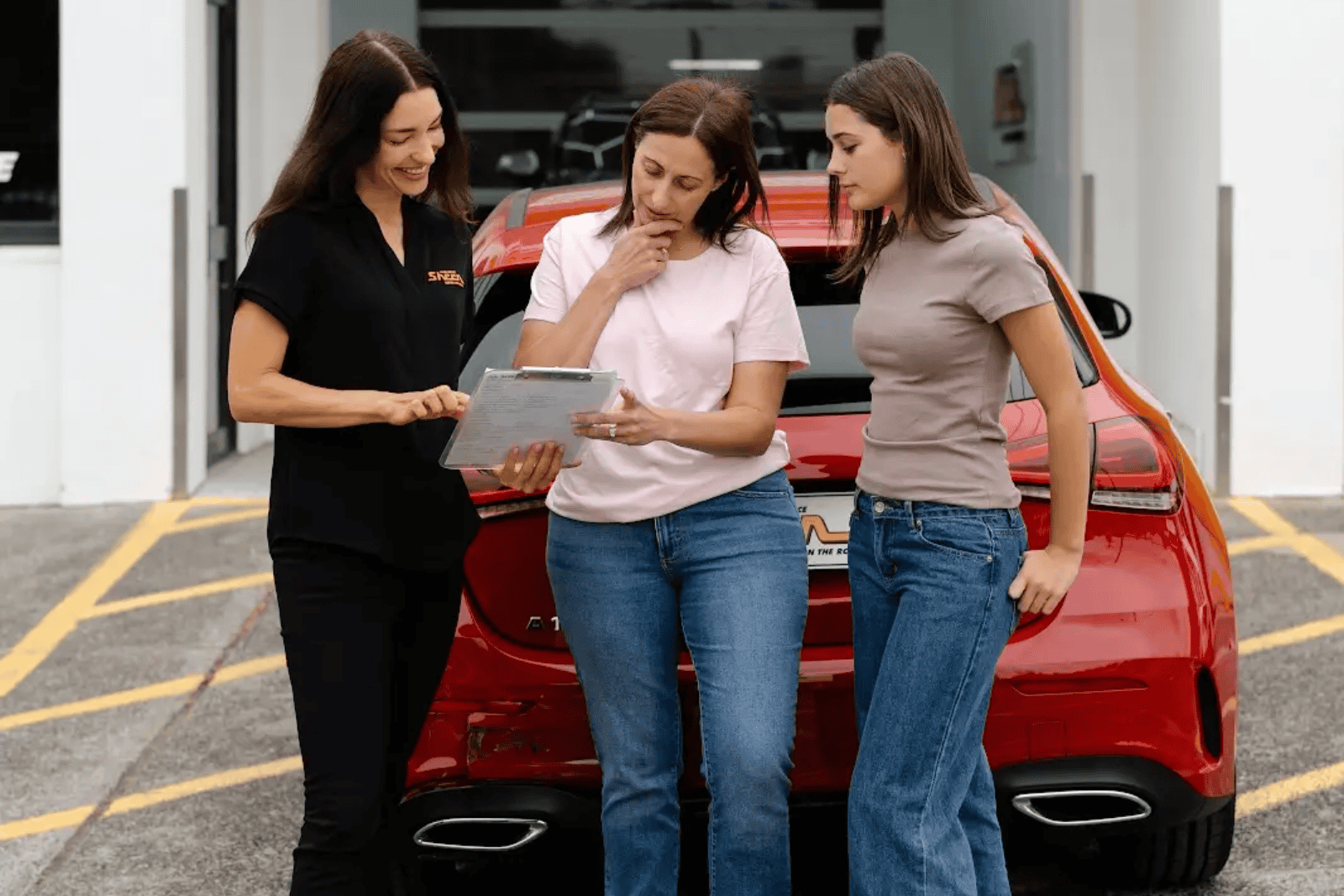 Sheen employee showing a clipboard to two women standing behind a red car with rear damage, outside an automotive repair centre.