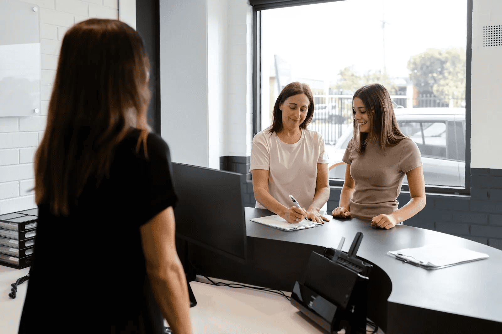 Two women smiling as one fills out paperwork at a front counter inside a Sheen repair centre, with a staff member standing across from them.