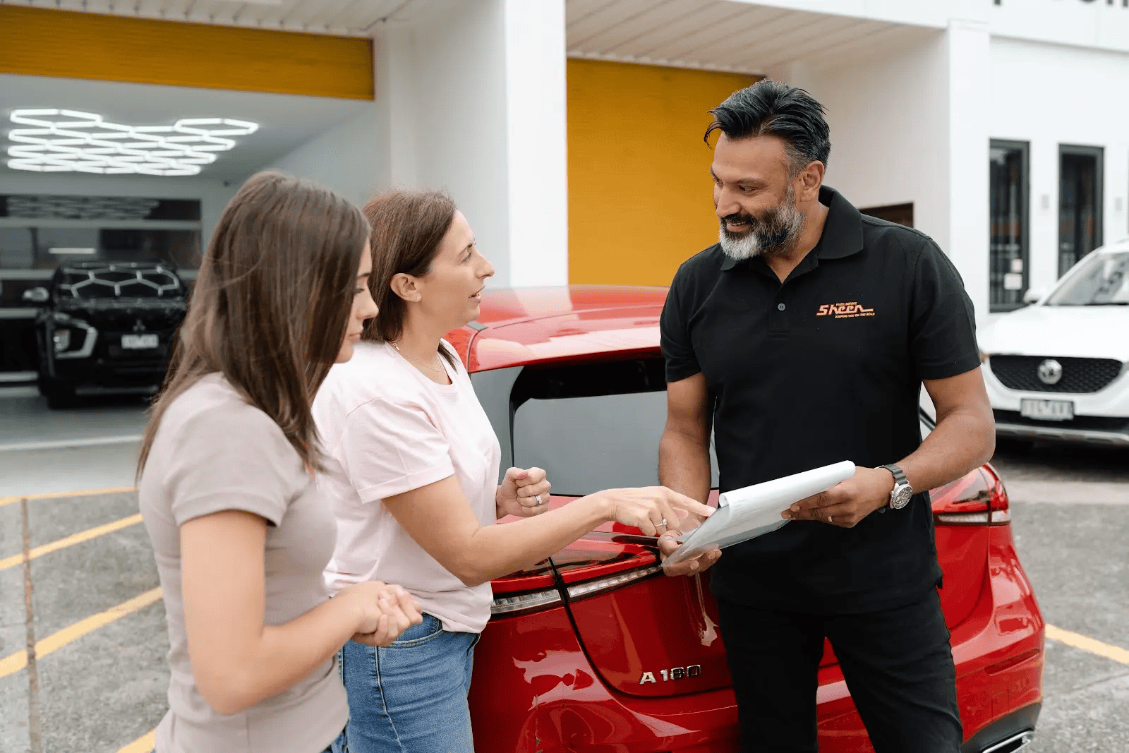 Sheen team member discussing paperwork with two women standing beside a red Mercedes A180 with rear damage, outside a repair centre.