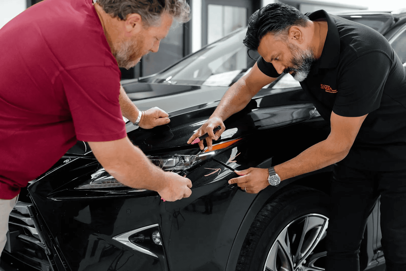 Sheen panel beater and technician inspecting and marking scratches on the front of a black vehicle inside a repair workshop.