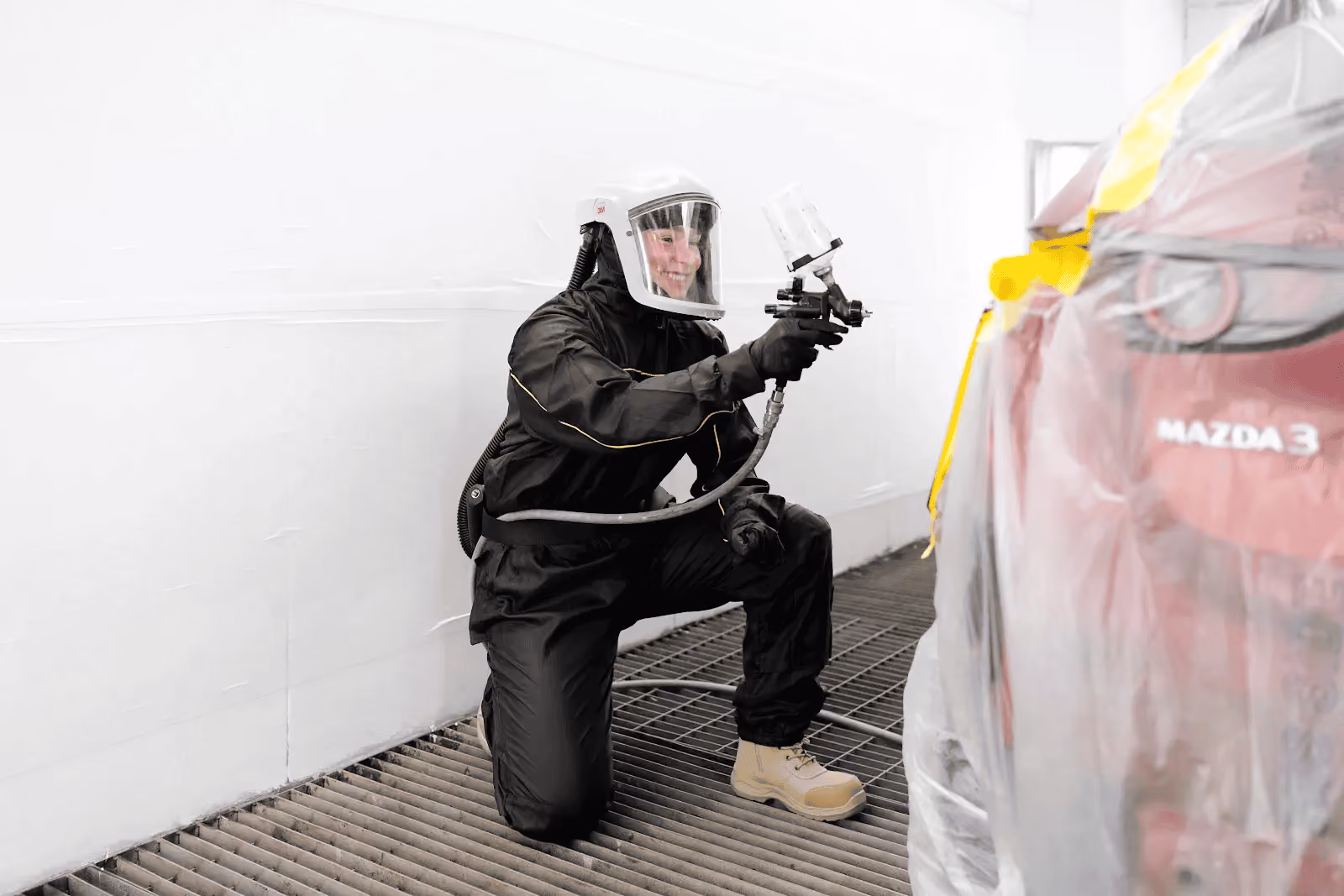 Sheen spray painter in protective gear applying paint to a red Mazda inside a controlled spray booth.