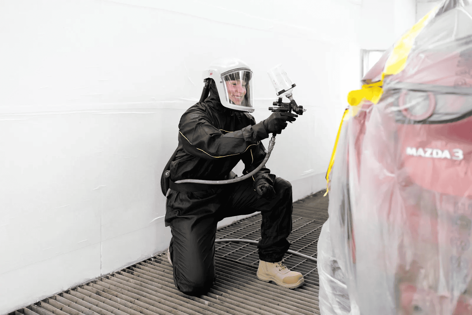 Sheen spray painter in protective gear applying paint to a red Mazda inside a controlled spray booth.