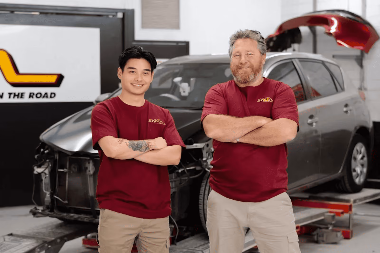 Two Sheen Panel Service technicians standing in front of a damaged vehicle inside a repair workshop.