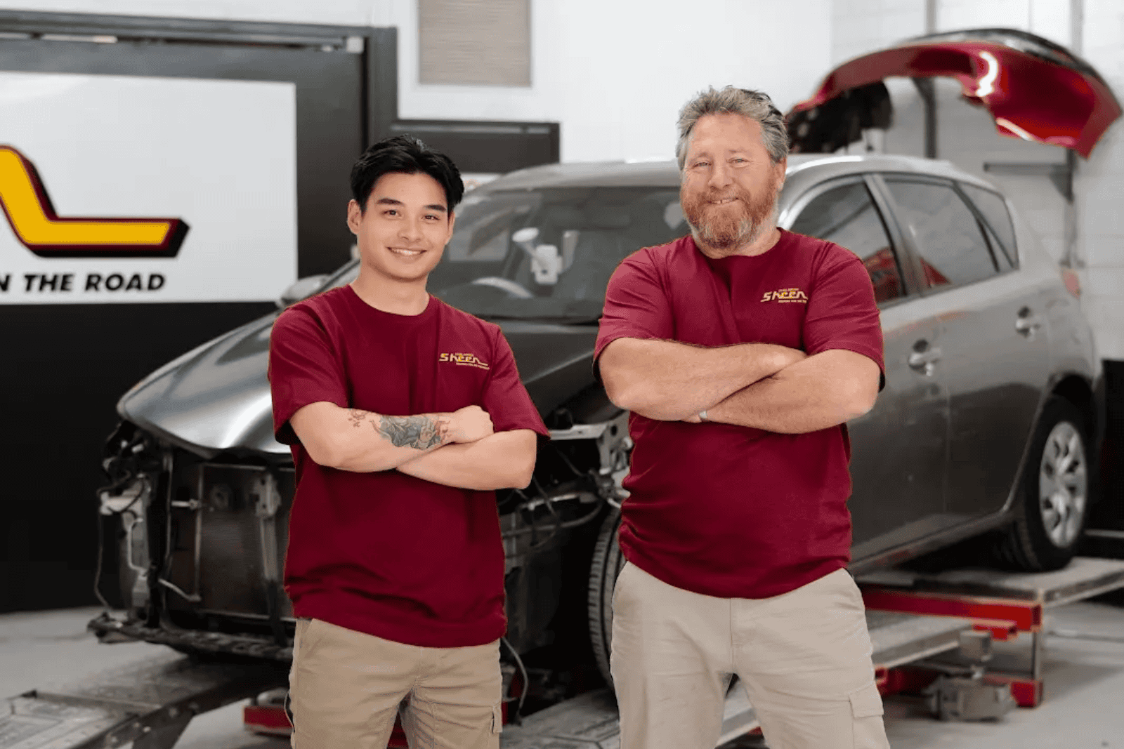 Two Sheen Panel Service technicians standing in front of a damaged vehicle inside a repair workshop.