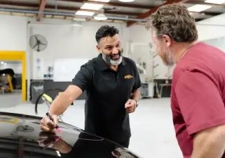 Sheen panel beater in Melbourne discussing repair costs with a customer while marking dents on a car bonnet inside a workshop.