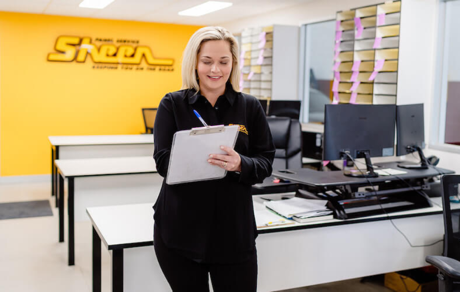 A smiling panel repair receptionist reviewing paperwork at a Sheen Group office desk, preparing accident claim details for a customer.