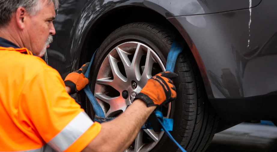 Sheen tow truck operator securing a car wheel with a strap during vehicle loading for towing.
