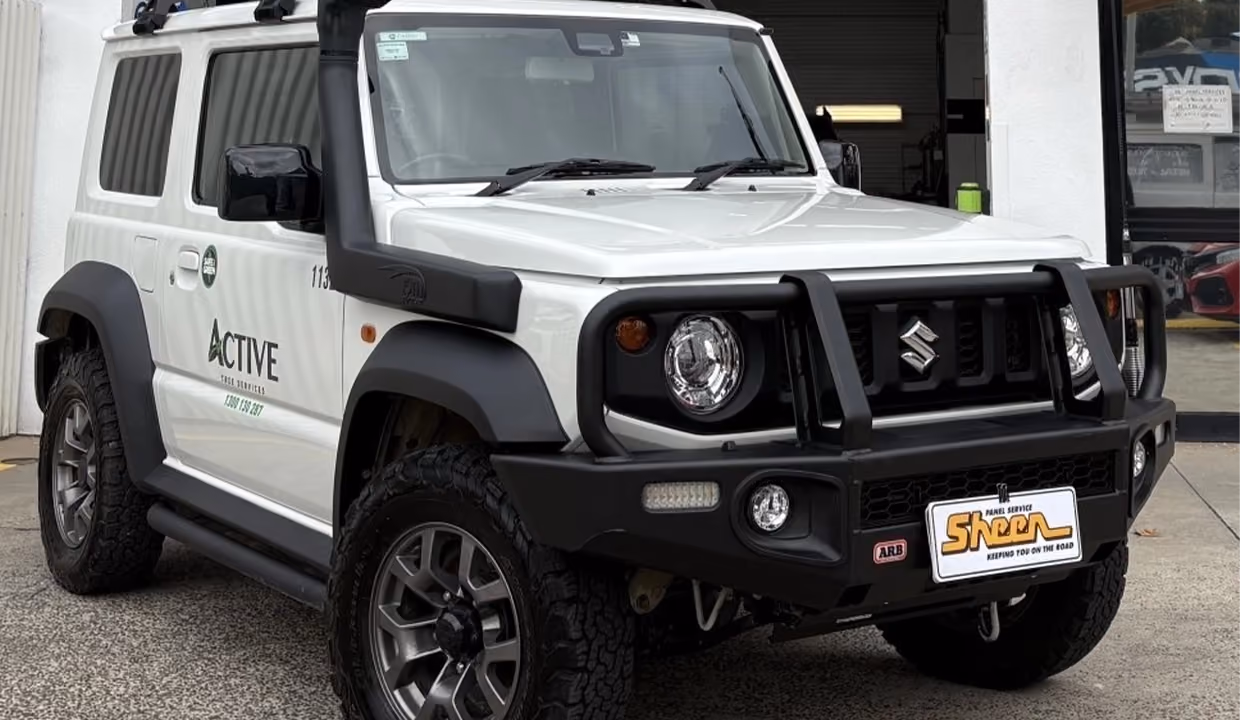 Front view of a white Suzuki Jimny after repair, with a straight bonnet, aligned grille, and fully restored front end parked outside a workshop.