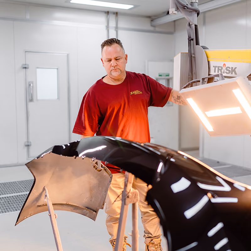 Sheen technician inspecting a repaired bumper under workshop lights.