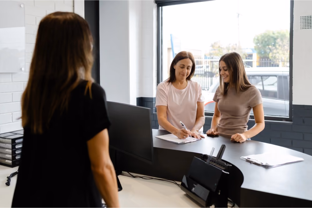 Two women filling out insurance paperwork at a service desk while a staff member looks on.