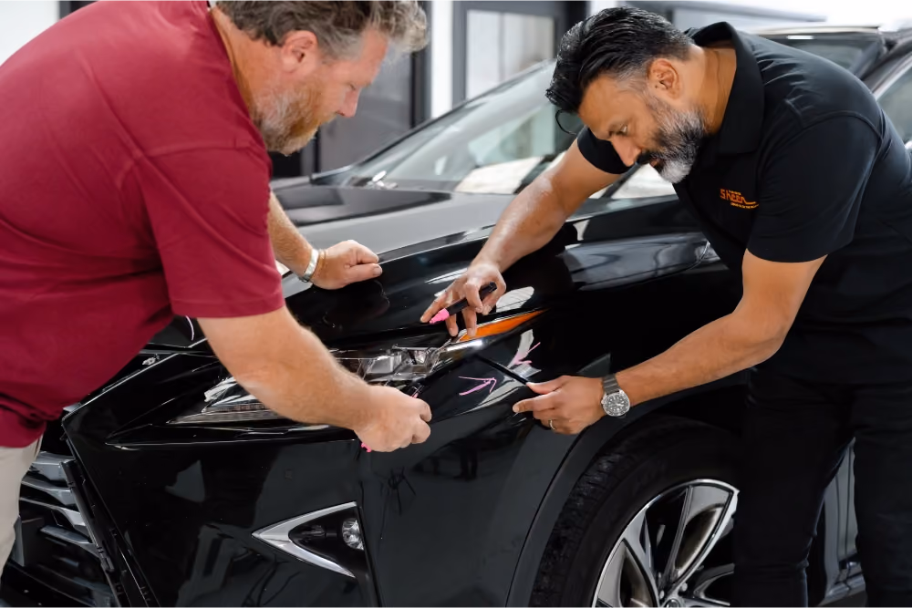 Two Sheen Group team members inspecting and marking damage on the front of a black car using pink markers in a repair workshop.