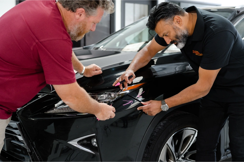 Two Sheen Group team members inspecting and marking damage on the front of a black car using pink markers in a repair workshop.