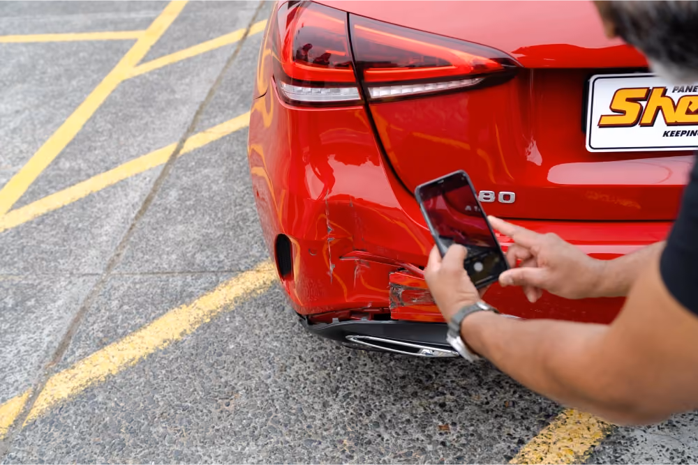 Person taking a photo of rear-end damage on a red car using a smartphone, showing a dent and scrapes near the bumper.]