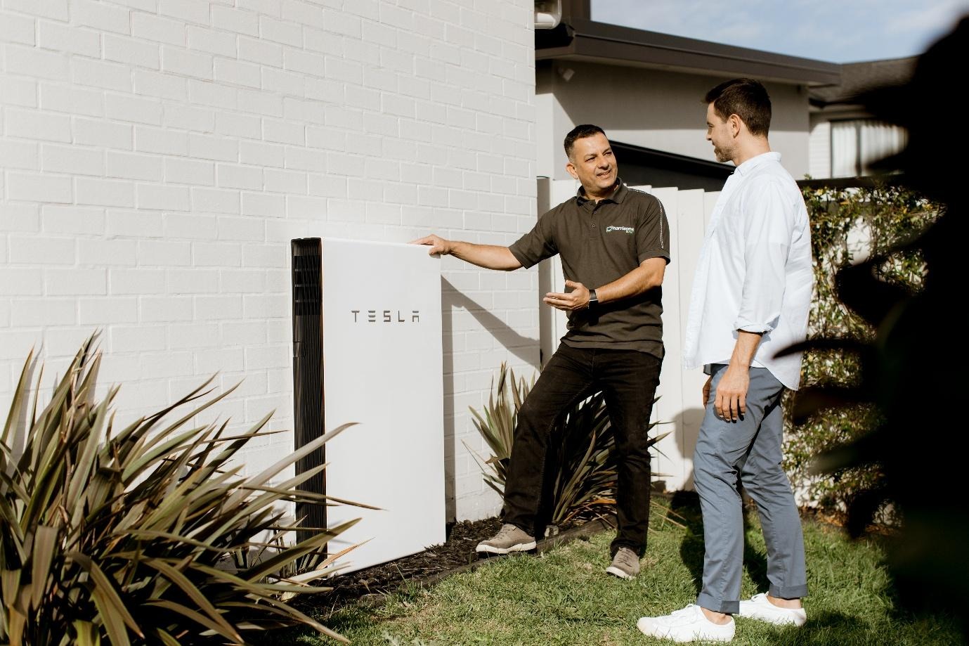 The image shows two people standing outside a Tesla electric car, examining the solar panel installation on their house.AI-generated content may be incorrect.