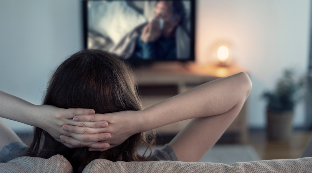 Back of a woman's head with her arms crossed behind her head watching TV