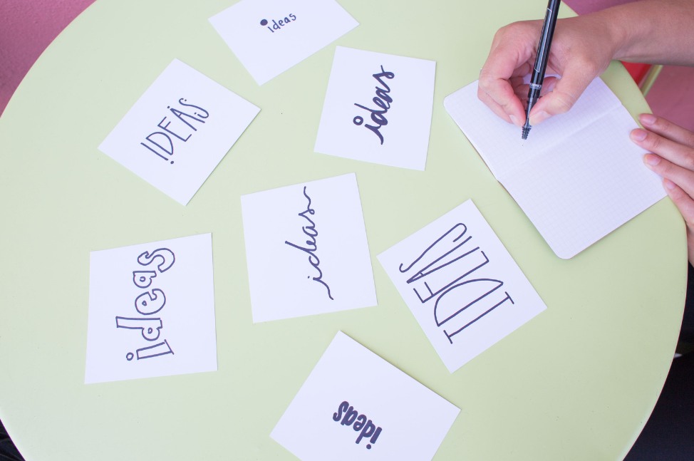 Several sticky notes on a green table. The word 'ideas' is written on every sticky note with different fonts. A person is writing something on a notepad with a black pen.
