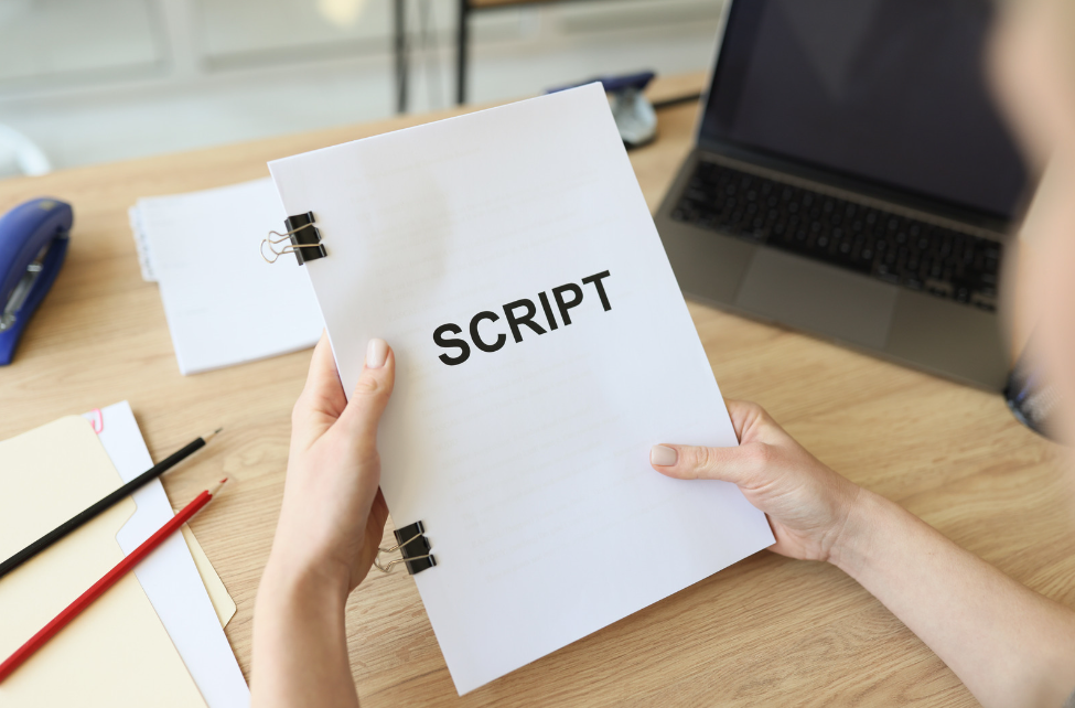 A person holding a script. There are colored pencils, a laptop, some paper, and a blue stapler on the desk.