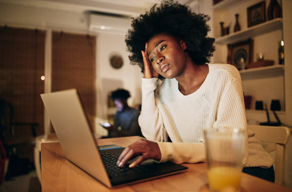 A woman is struggling with her project. She is resting her head on her right hand, looking at her laptop. There is a glass on the table.