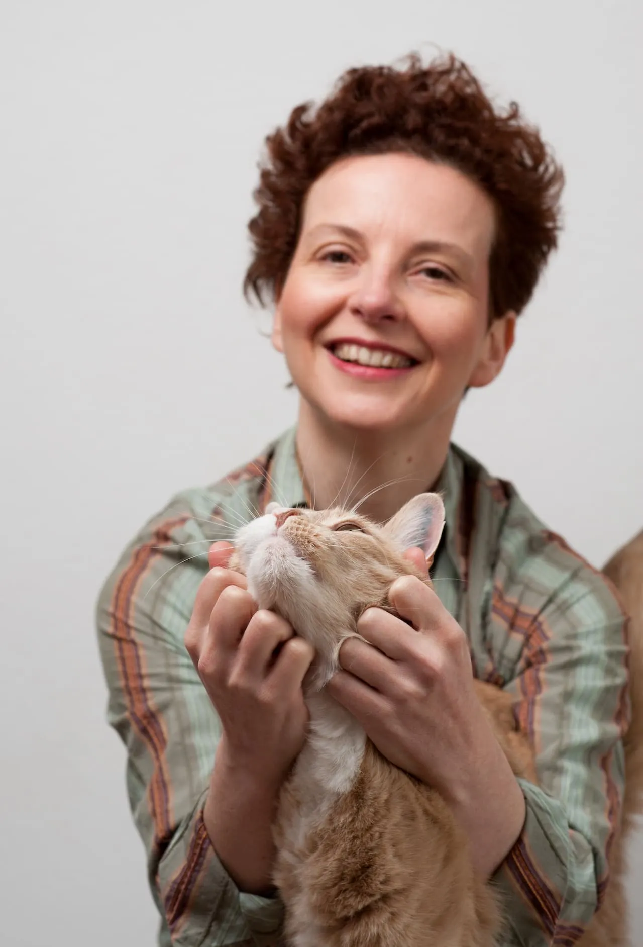 Smiling woman in a striped shirt gently holding a content orange and white cat under its chin.