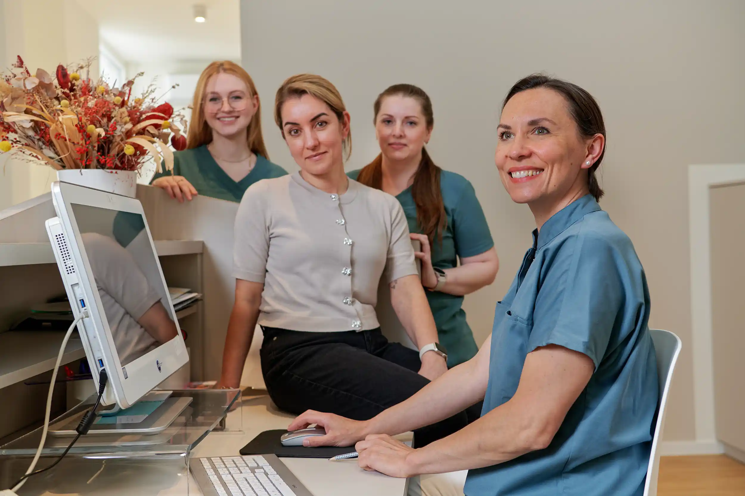 Team: Four women in a reception area, one seated at a computer desk smiling and the others standing behind her, with a bouquet of dried flowers on the counter.