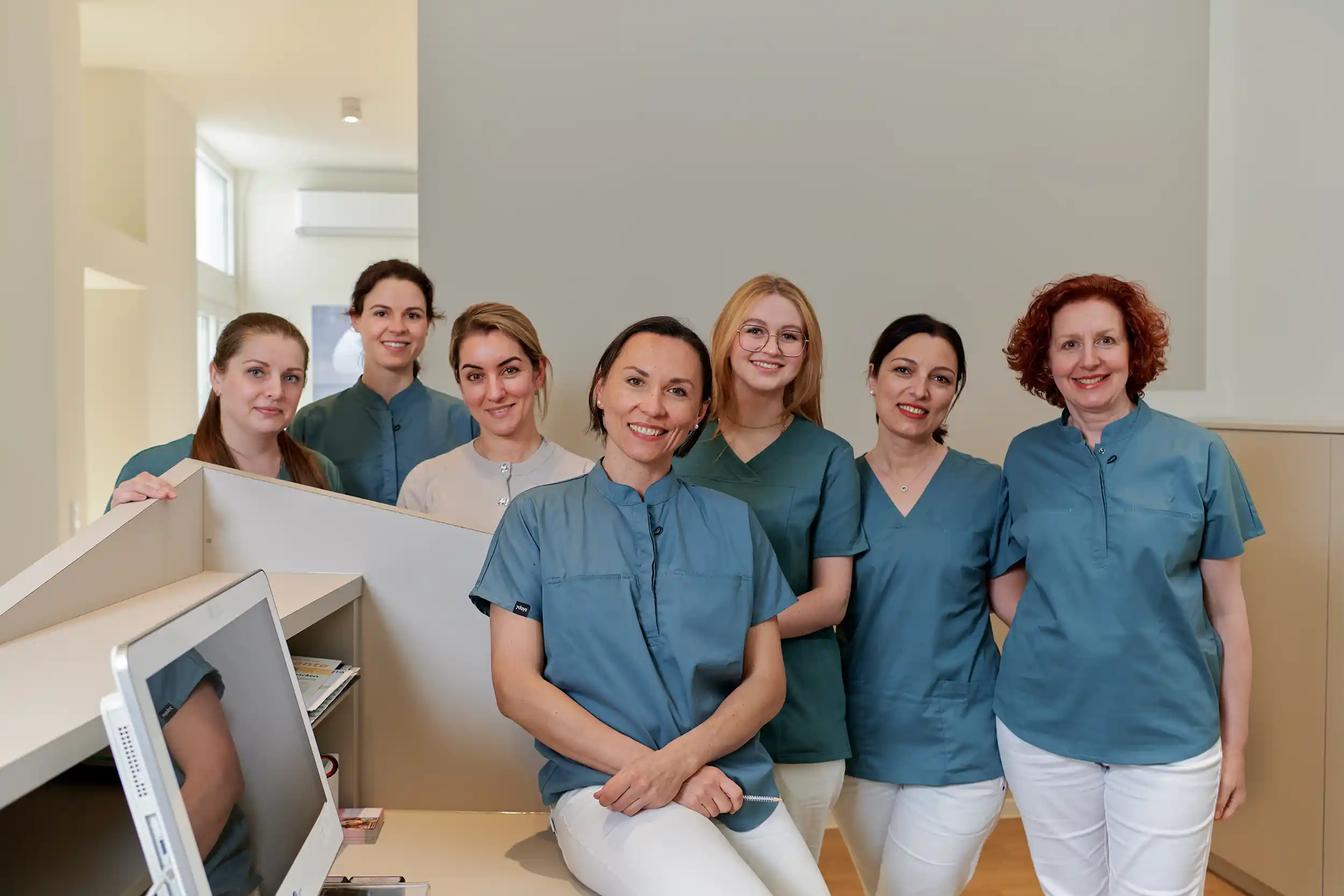Team: Seven women in medical scrubs and white pants smiling at a reception desk in a clinical setting.