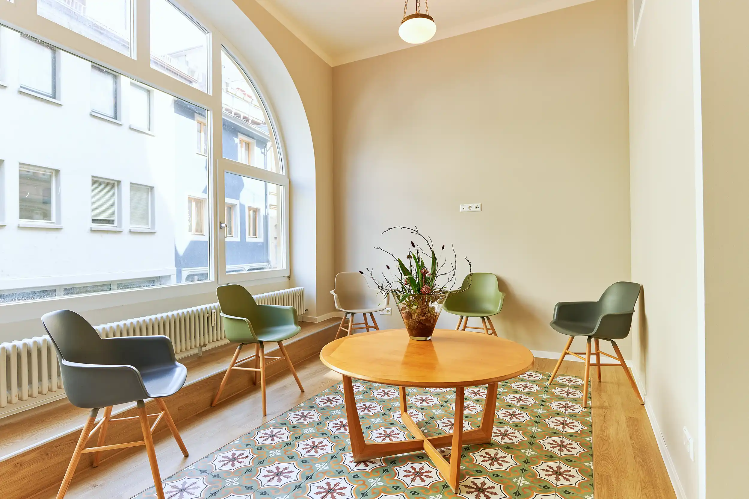 Bright waiting room with an arched window, five modern armchairs, a round wooden table with a floral centerpiece, and decorative tiled flooring.