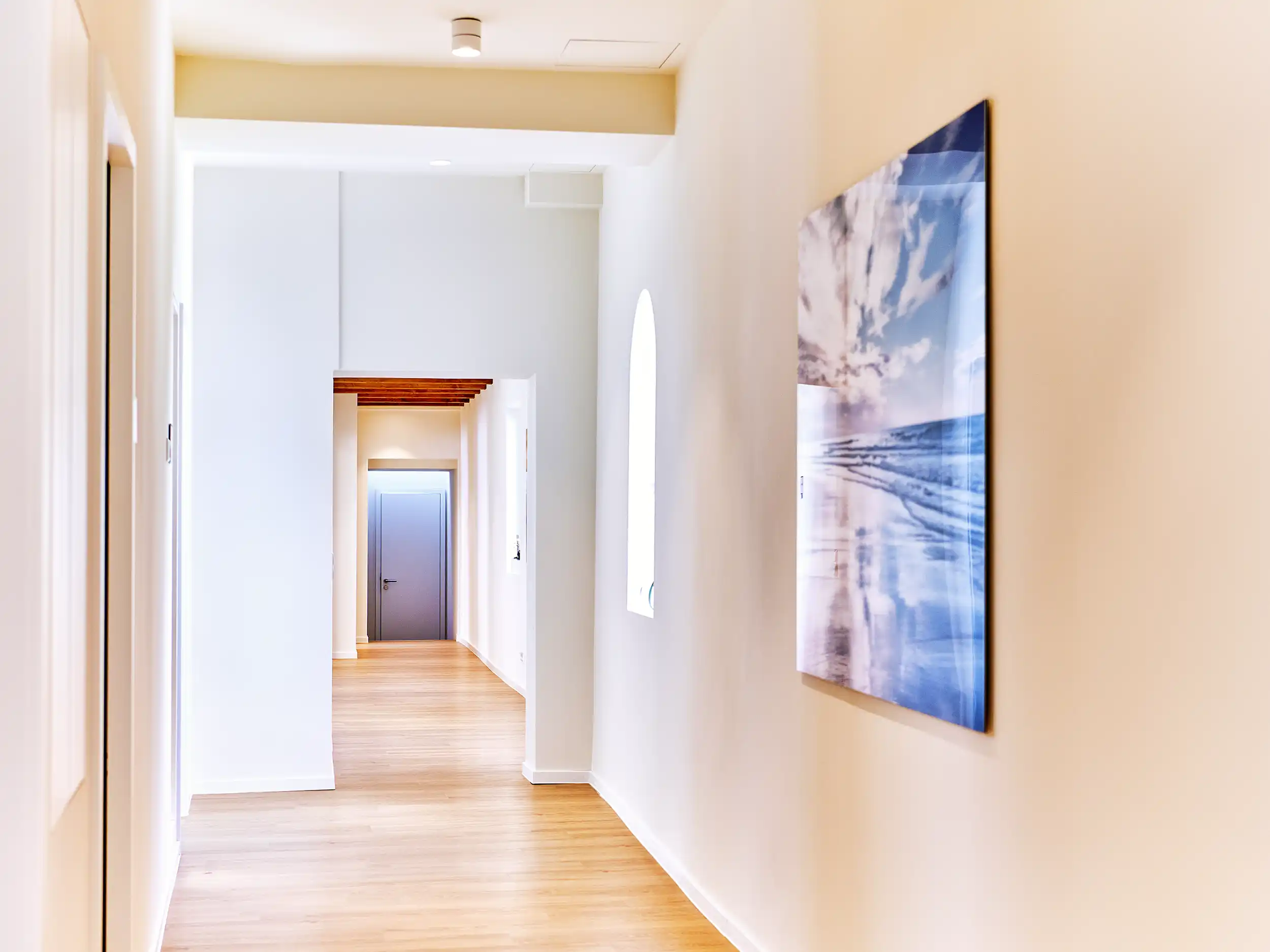 Bright hallway with light wooden floor, white walls, arched window, wall art of a cloudy sky, and a grey door at the end.