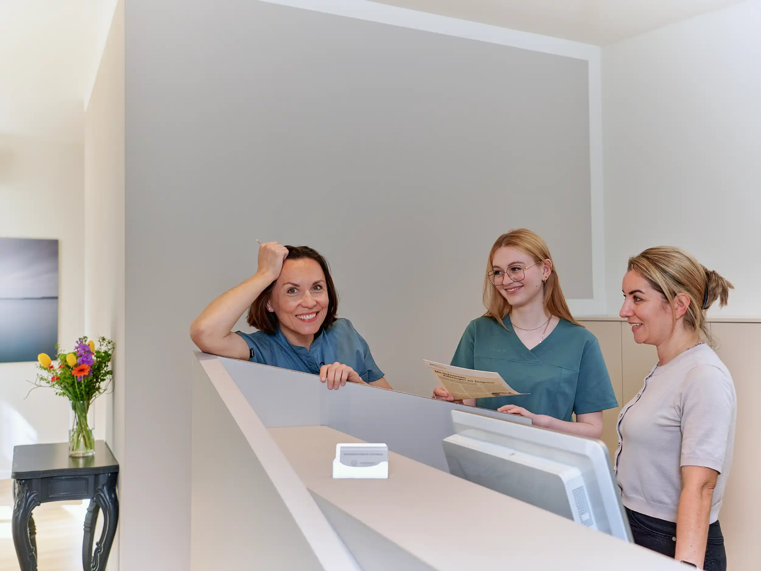 Three women smiling at a reception desk in a modern office with a vase of colorful flowers on a side table.