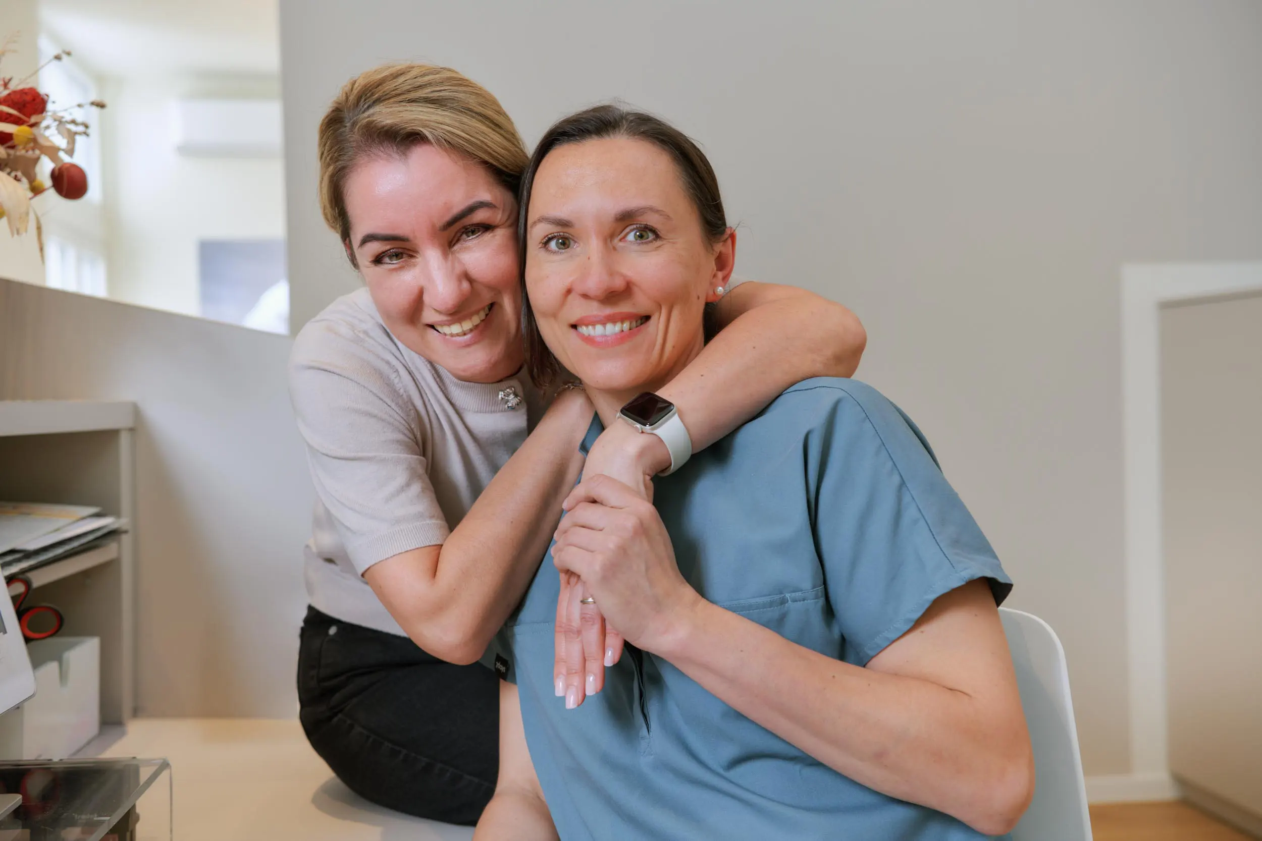 Frau Stella und Frau Dr. Heeren: Two smiling women sitting closely indoors, one wearing a blue scrub top with an Apple Watch, the other hugging her from behind.