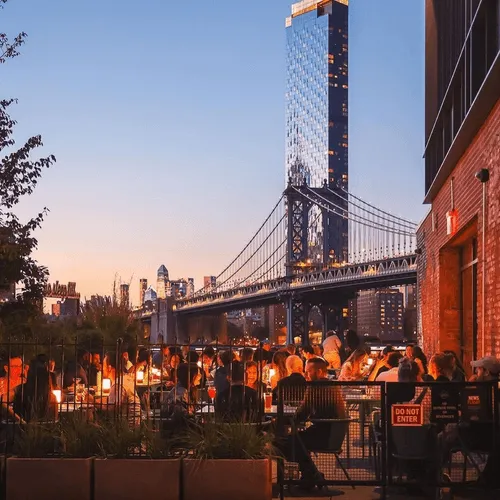 People sitting outside at a restaurant patio overlooking the river with suspension bridge and New York skyline in the background.