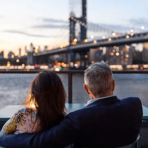 A couple sits on a bench looking at the river, suspension bridge and New York Skyline.