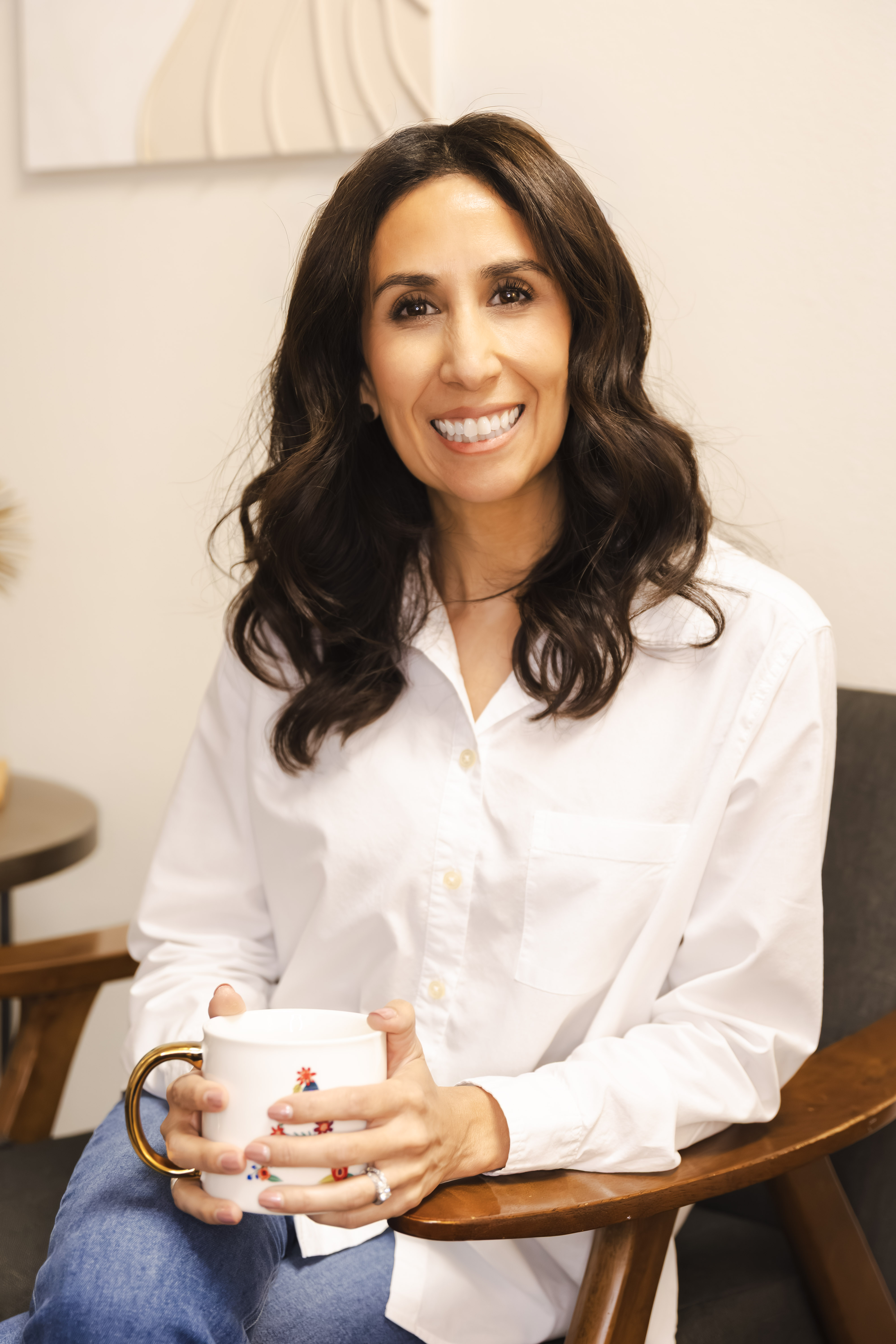 Smiling woman with wavy dark hair wearing a white shirt and blue jeans, sitting in a wooden chair and holding a white mug with a gold handle.