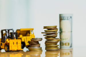 Mini construction vehicle models next to piles of coins and $100 bills. 