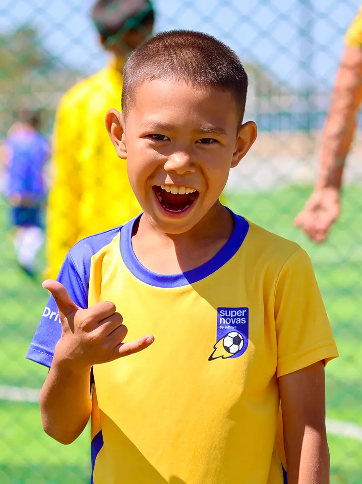 Smiling boy in yellow and blue soccer jersey making a shaka hand sign on a soccer field.
