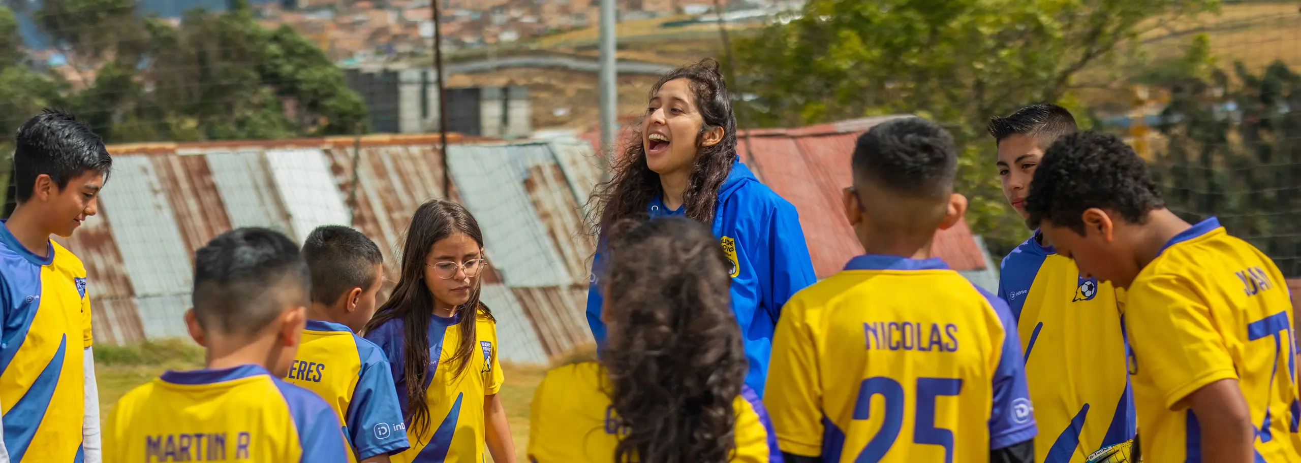 Youth soccer coach enthusiastically speaking to a group of children in yellow and blue jerseys gathered on a field.
