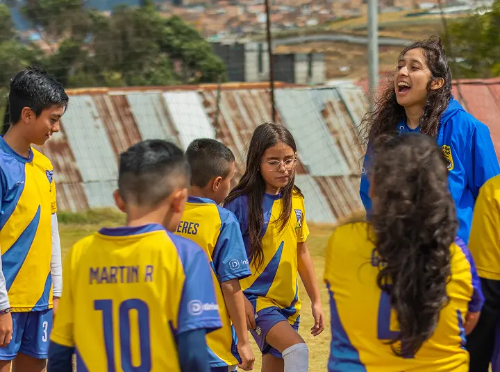 Group of young soccer players in yellow and blue uniforms gathered on a field with a female coach in a blue jacket interacting with them.