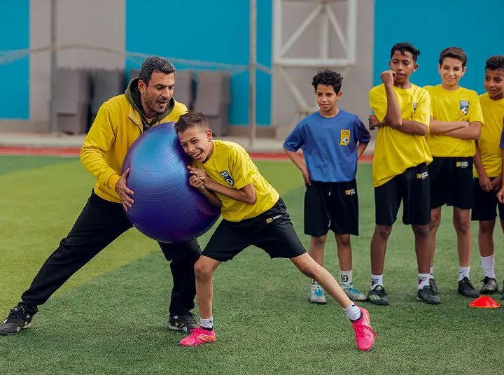 Coach assisting a boy in yellow shirt pushing a large purple exercise ball on a sports field while other boys watch.