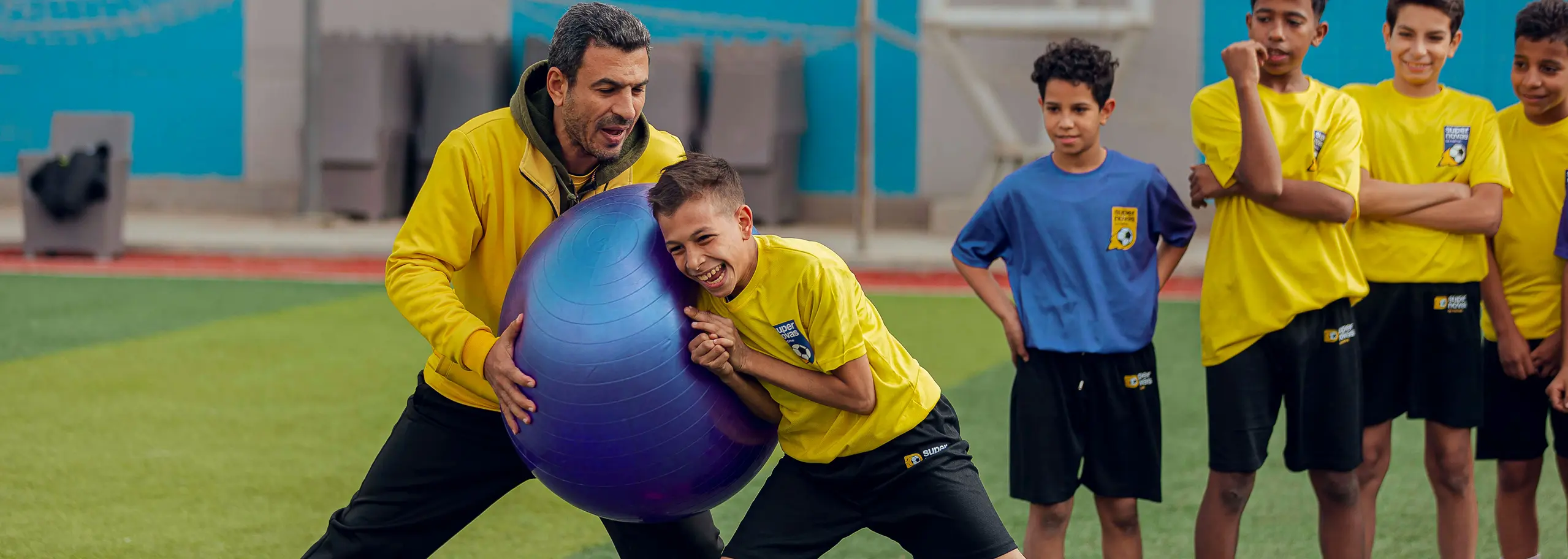 Coach in yellow jacket and boy in yellow shirt holding a large purple exercise ball, with other children in yellow and blue sports uniforms watching on a green field.