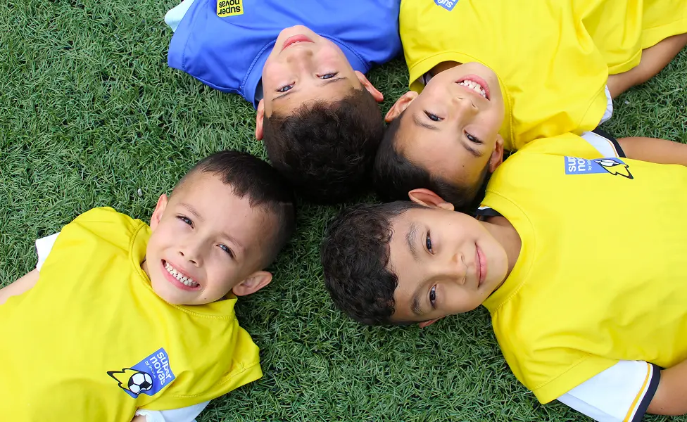 Four young boys lying on green grass in yellow and blue sports jerseys smiling at the camera.
