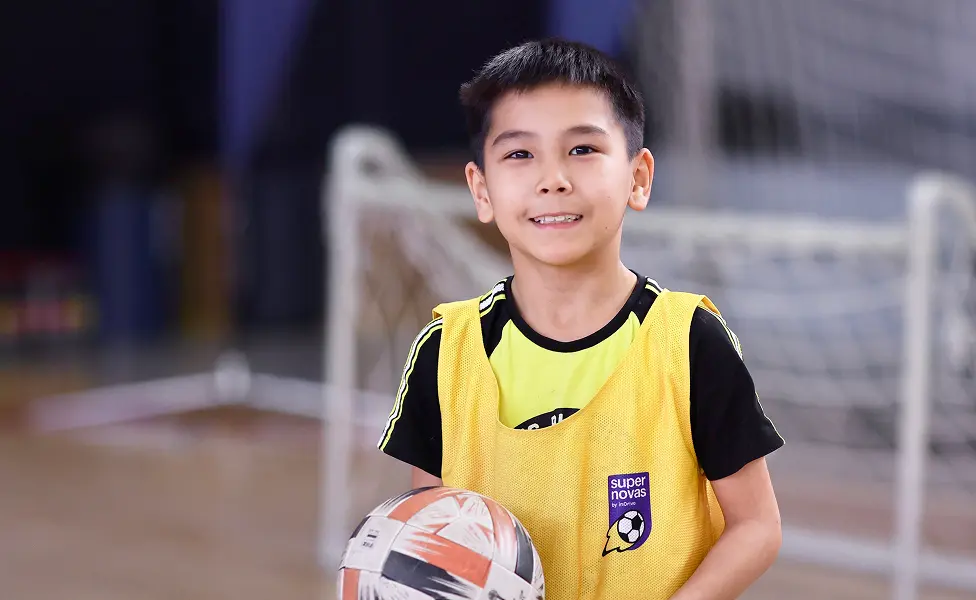 Young boy holding a soccer ball indoors, wearing a yellow sports vest with a soccer goal in the background.