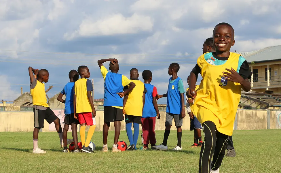 A group of children wearing yellow and blue vests playing soccer on a grassy field, one boy in a yellow vest smiling and running toward the camera.