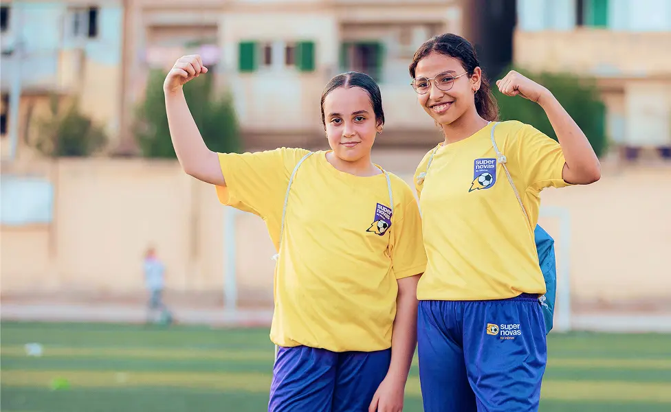 Two girls in yellow sports shirts and blue shorts flexing their arms on a soccer field.